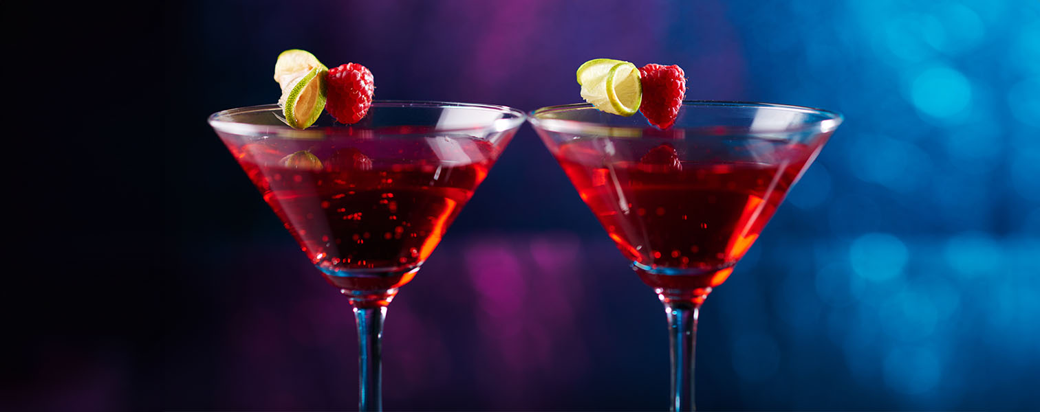 Two red cocktails with a raspberry and rolled cucumber slice on the rim. The background is a purple blue ombre.