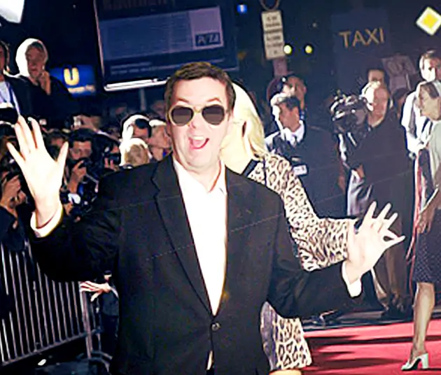 An image of Mike Colvin in a black suit jacket, white button up and gold aviator sunglasses on a red carpet.